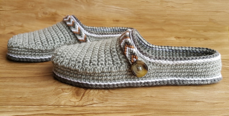 Peut inclure: Une paire de pantoufles en crochet gris avec des bordures blanches et brunes. Les pantoufles ont un bouton sur le dessus et le texte "Crochet Oasis" est visible sur la pantoufle de droite.