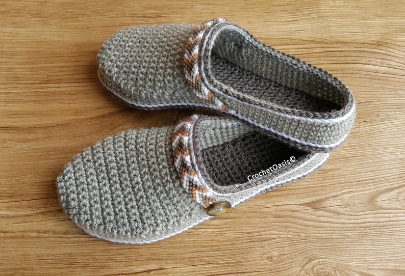 Peut inclure: Une paire de pantoufles en crochet gris avec des bordures blanches et orange. Les pantoufles ont un bouton sur le dessus et le texte "CrochetOasis" est visible sur le c&ocirc;t&eacute;.