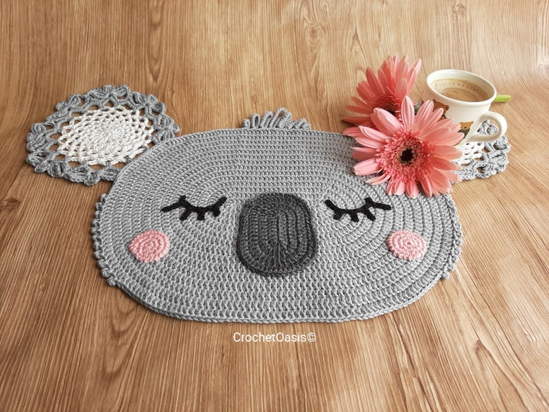 May include: A gray crocheted koala-shaped coaster with pink and black details. The coaster is on a wooden surface with two pink flowers and a coffee cup.