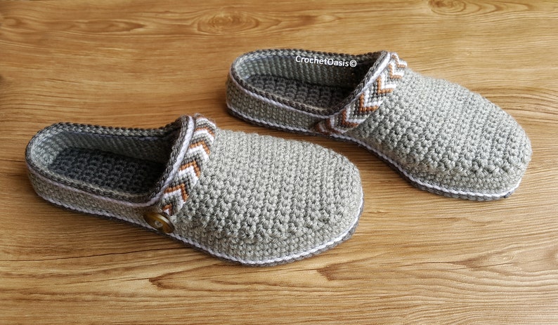 Peut inclure: Une paire de pantoufles en crochet gris avec un motif chevron blanc et marron. Les pantoufles ont un d&eacute;tail bouton et sont faites avec un fil doux.