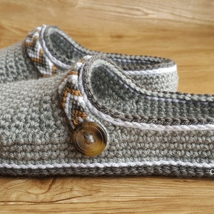 Peut inclure: Une paire de pantoufles en crochet gris avec une bordure blanche et un bouton marron. Les pantoufles ont un design textur&eacute; et sont faites avec un fil doux.