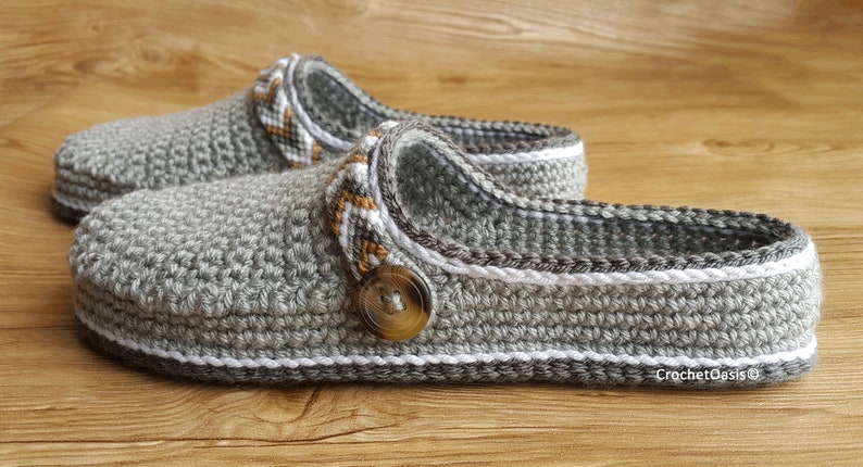 Peut inclure: Une paire de pantoufles en crochet gris avec une bordure blanche et un bouton marron. Les pantoufles ont un design textur&eacute; et sont faites avec un fil doux.