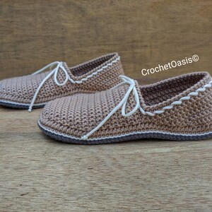 May include: A pair of crocheted brown slippers with white laces and a white crocheted trim. The slippers have a brown sole and are sitting on a wooden surface. CrochetOasis&reg; is written in white text on the right side of the image.