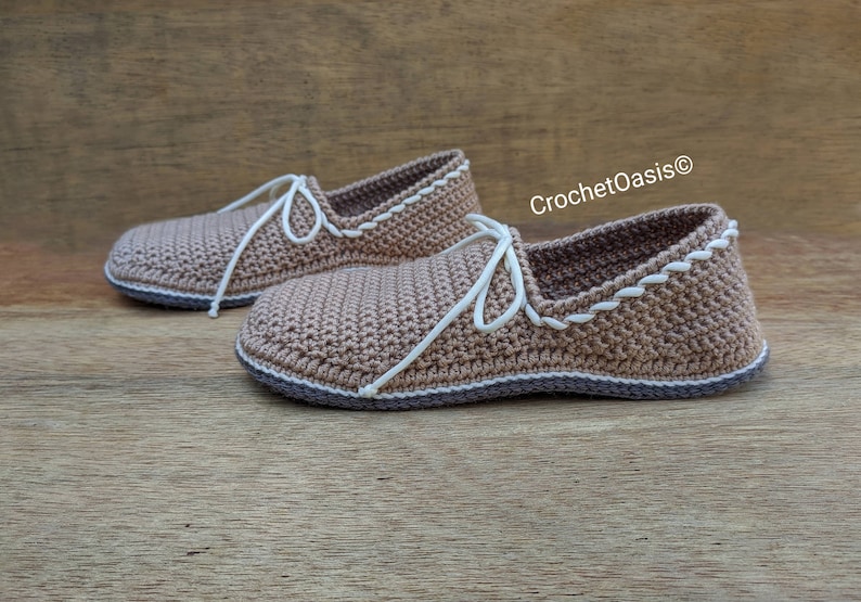 May include: A pair of crocheted brown slippers with white laces and a white crocheted trim. The slippers have a brown sole and are sitting on a wooden surface. CrochetOasis&reg; is written in white text on the right side of the image.