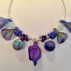 Puede incluir: Un collar morado y azul con una variedad de cuentas y colgantes. El collar presenta un gran colgante morado en el centro, rodeado de cuentas y colgantes más pequeños.