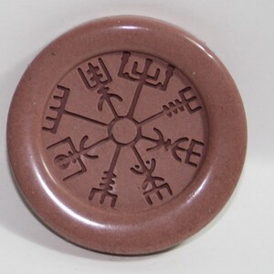Può includere: Una piastrella di ceramica circolare marrone con un design in rilievo di una rosa dei venti. La rosa dei venti è un simbolo di direzione e guida.