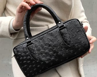 May include: Black rectangular handbag with a textured finish and a top handle. The bag features a zip closure and silver hardware. The bag is held by a person wearing a cream-coloured coat.
