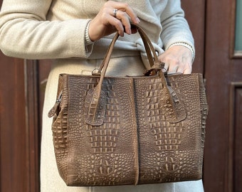 May include: A brown leather tote bag with a crocodile embossed pattern. The bag has two top handles and a zipper closure.