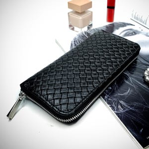 Puede incluir: Cartera de cuero negro tejido con cremallera plateada. La cartera es rectangular y tiene un patrón texturizado. Un frasco de esmalte de uñas, un tubo de lápiz labial rojo y una revista también están en la imagen.