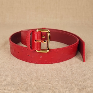 Op de afbeelding: Een felrode riem met een getextureerd oppervlak met kleine, gelijkmatig verdeelde inkepingen. De riem is voorzien van een rechthoekige goudkleurige metalen gesp en een bijpassende rode leren lus om het uiteinde vast te zetten.