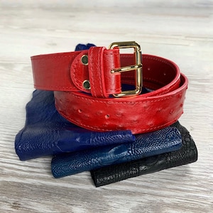 Puede incluir: Un cinturón de cuero rojo con una hebilla dorada y un patrón de avestruz texturizado. El cinturón está enrollado y se encuentra encima de una pila de cuero azul y negro.