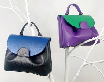 May include: Two leather handbags with contrasting colored top flaps. The handbag on the left has a black bottom and a blue top flap. The handbag on the right has a purple bottom and a green top flap.