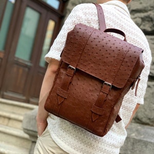 May include: A brown leather backpack with a textured ostrich pattern. The backpack has two adjustable buckles on the front flap and a zipper closure.