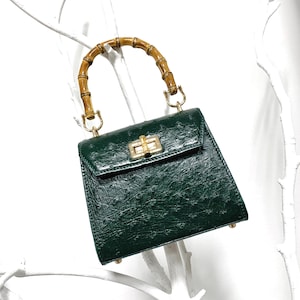 May include: A dark green handbag with a bamboo handle and gold-coloured hardware. The bag has a textured surface and a rectangular shape with a flap closure. The handle is curved and made of light brown bamboo.