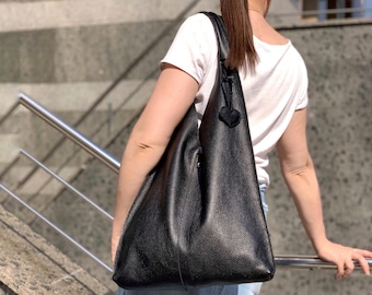 Puede incluir: Un bolso tote de cuero negro con forma triangular. El bolso tiene una sola correa y un pequeño charm en forma de corazón.