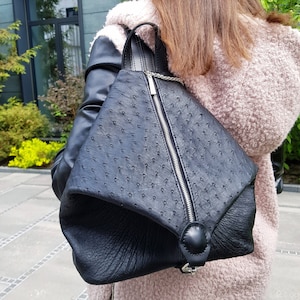 Puede incluir: Una mochila de cuero negro con forma triangular y un patrón de piel de avestruz en relieve. La mochila tiene una cremallera plateada y un detalle de cadena plateada.