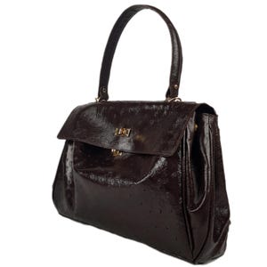 May include: A dark brown handbag with a glossy finish. The bag features a top handle, a flap closure with a gold-colored clasp, and a textured surface. The bag is designed with a structured shape and a single handle.