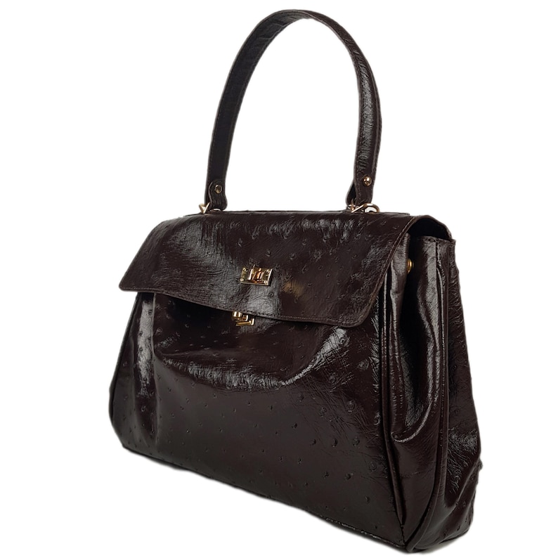 May include: A dark brown handbag with a glossy finish. The bag features a top handle, a flap closure with a gold-colored clasp, and a textured surface. The bag is designed with a structured shape and a single handle.