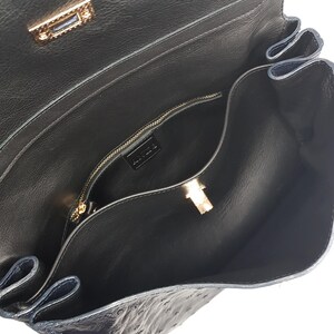 May include: Open view of a black leather handbag with a textured surface. The interior reveals a zippered pocket and a gold-colored clasp. The bag's design includes a flap closure and a gold-toned metal lock.