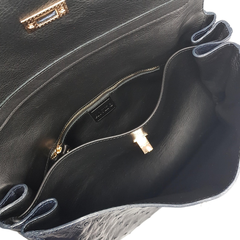 May include: Open view of a black leather handbag with a textured surface. The interior reveals a zippered pocket and a gold-colored clasp. The bag's design includes a flap closure and a gold-toned metal lock.