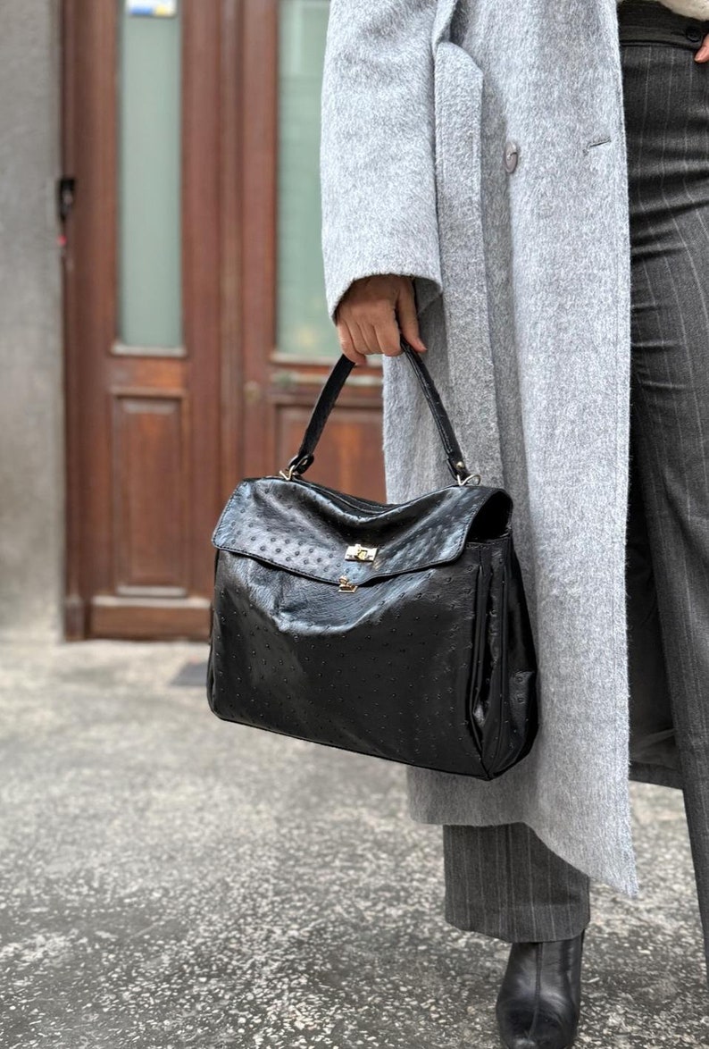 May include: Black leather handbag with a textured ostrich-like pattern. The bag has a flap closure with a gold-colored clasp and a top handle. The bag is held by a person wearing a gray coat and pinstripe pants.