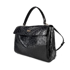 May include: Black leather handbag with a top handle and a flap closure. The bag features a textured surface with small, evenly spaced dots. A gold-colored clasp secures the flap. Includes a shoulder strap.