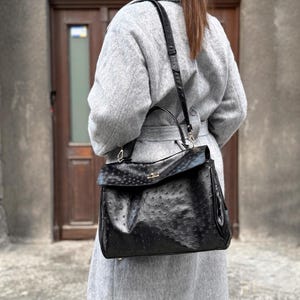 May include: A black leather handbag with a textured surface and a flap closure. The bag has a top handle and a detachable shoulder strap. The hardware is gold-toned. The bag is worn over a gray coat.