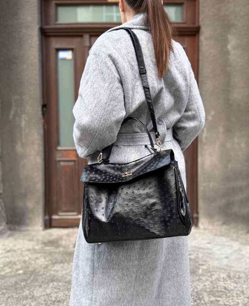 May include: A black leather handbag with a textured surface and a flap closure. The bag has a top handle and a detachable shoulder strap. The hardware is gold-toned. The bag is worn over a gray coat.