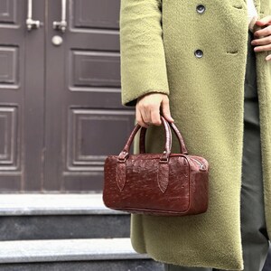 May include: A brown leather handbag with a textured surface and two handles. The bag has a rectangular shape and a zipper closure. The person is wearing a long olive green coat.