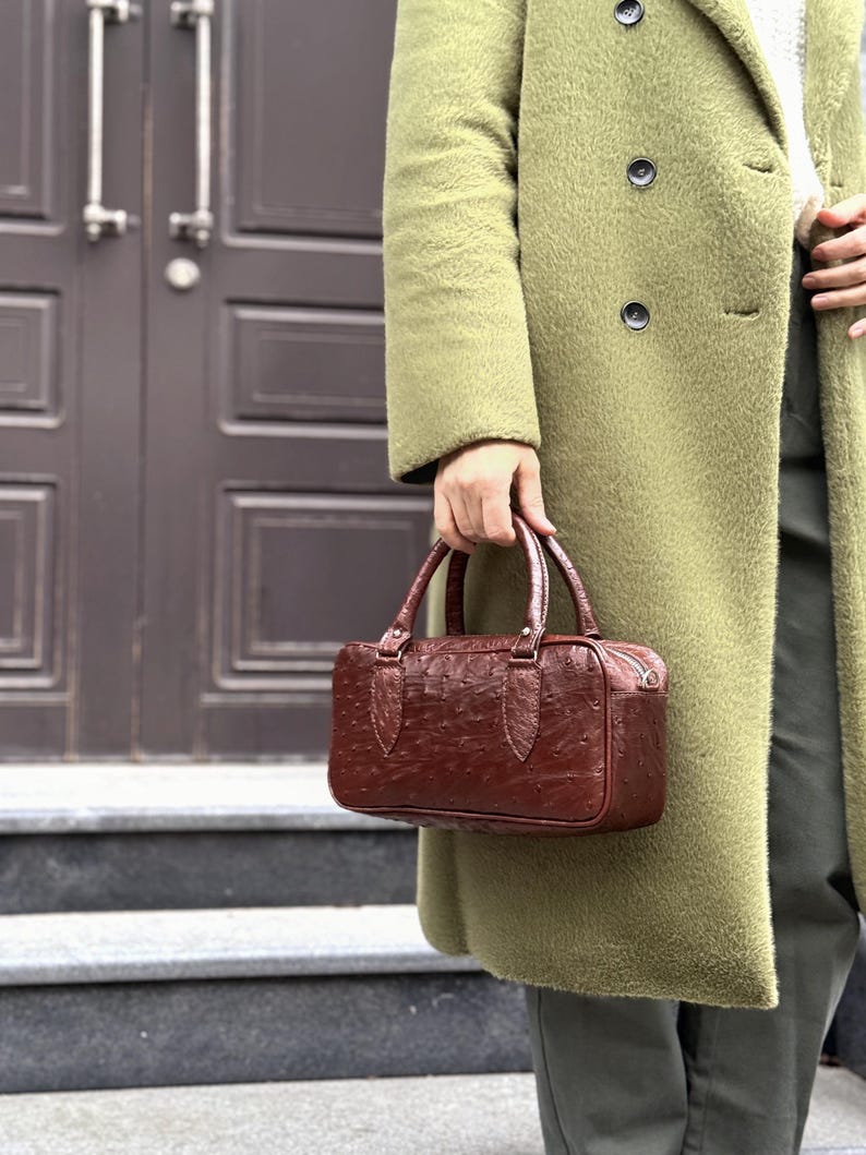 May include: A brown leather handbag with a textured surface and two handles. The bag has a rectangular shape and a zipper closure. The person is wearing a long olive green coat.