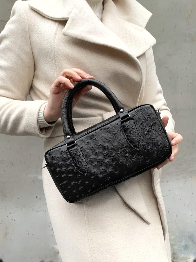 May include: Black rectangular handbag with a textured surface and a top handle. The bag has a zipper closure and silver-toned hardware. The bag is held by a person wearing a cream-colored coat.