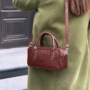 May include: A brown leather handbag with a textured surface and a top handle. The bag has a rectangular shape and a detachable shoulder strap. The bag is worn over a green coat.