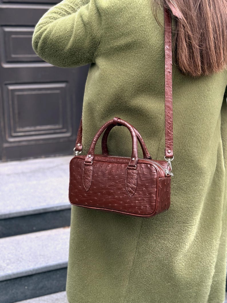 May include: A brown leather handbag with a textured surface and a top handle. The bag has a rectangular shape and a detachable shoulder strap. The bag is worn over a green coat.
