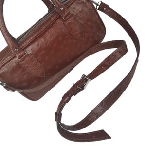 May include: A brown leather handbag with a textured surface and a silver zipper. The bag has a matching adjustable shoulder strap and a top handle. The bag is approximately 10 inches long.