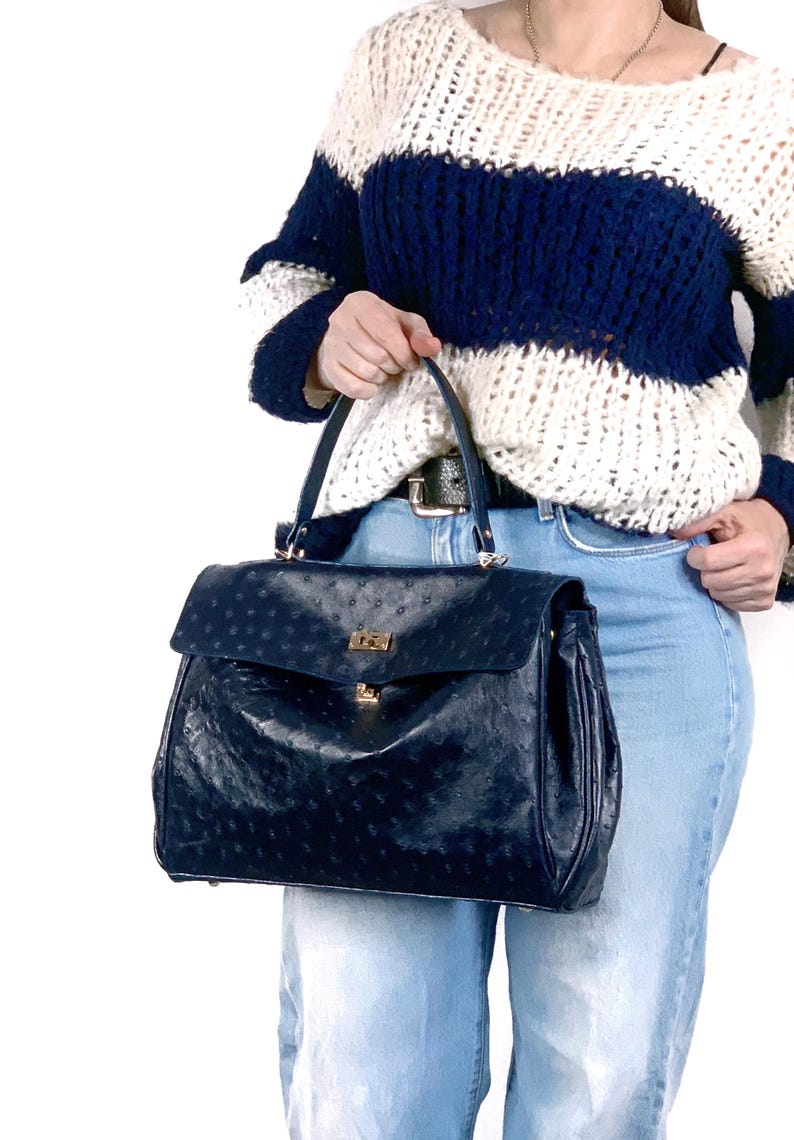 May include: A navy blue handbag with a flap closure and gold-colored hardware. The bag has a textured surface and a top handle. The bag is held by a person wearing a white and navy blue striped sweater and blue jeans.