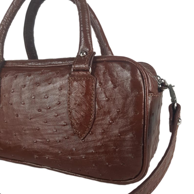 May include: A brown leather handbag with a textured surface and a small handle. The bag has a zipper closure and a detachable shoulder strap. The bag is a dark brown color.