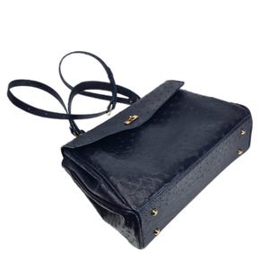 May include: A navy blue handbag with a textured ostrich leather design. The bag features a flap closure with a gold-colored clasp and a long, adjustable strap. Gold-colored studs are on the bottom of the bag.