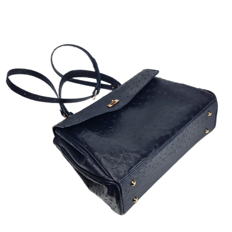 May include: A navy blue handbag with a textured ostrich leather design. The bag features a flap closure with a gold-colored clasp and a long, adjustable strap. Gold-colored studs are on the bottom of the bag.