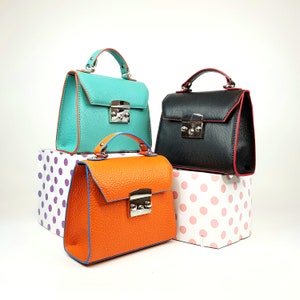 May include: Three small leather handbags in different colors: teal, black, and orange. Each bag has a silver clasp and a top handle. The bags are arranged on a white surface with polka dot patterned boxes.