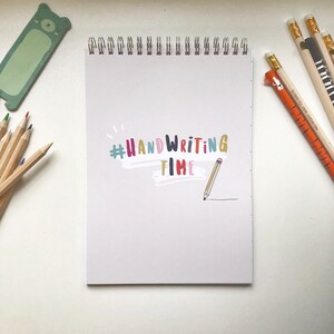 Może przedstawiać: Biały notatnik w spiralnej oprawie z napisem "#Handwriting Time" w kolorowych literach, wraz z ilustracją ołówka. Na zdjęciu znajdują się również kredki, ołówki z motywami zwierzęcymi i zakładka w kształcie misia.