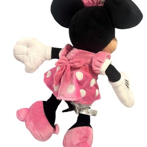 May include: A pink and white polka dot dress with a white bow on the back. The dress is worn by a plush toy of Minnie Mouse, who has black ears and a black bow on her head. The toy is standing on a white background.