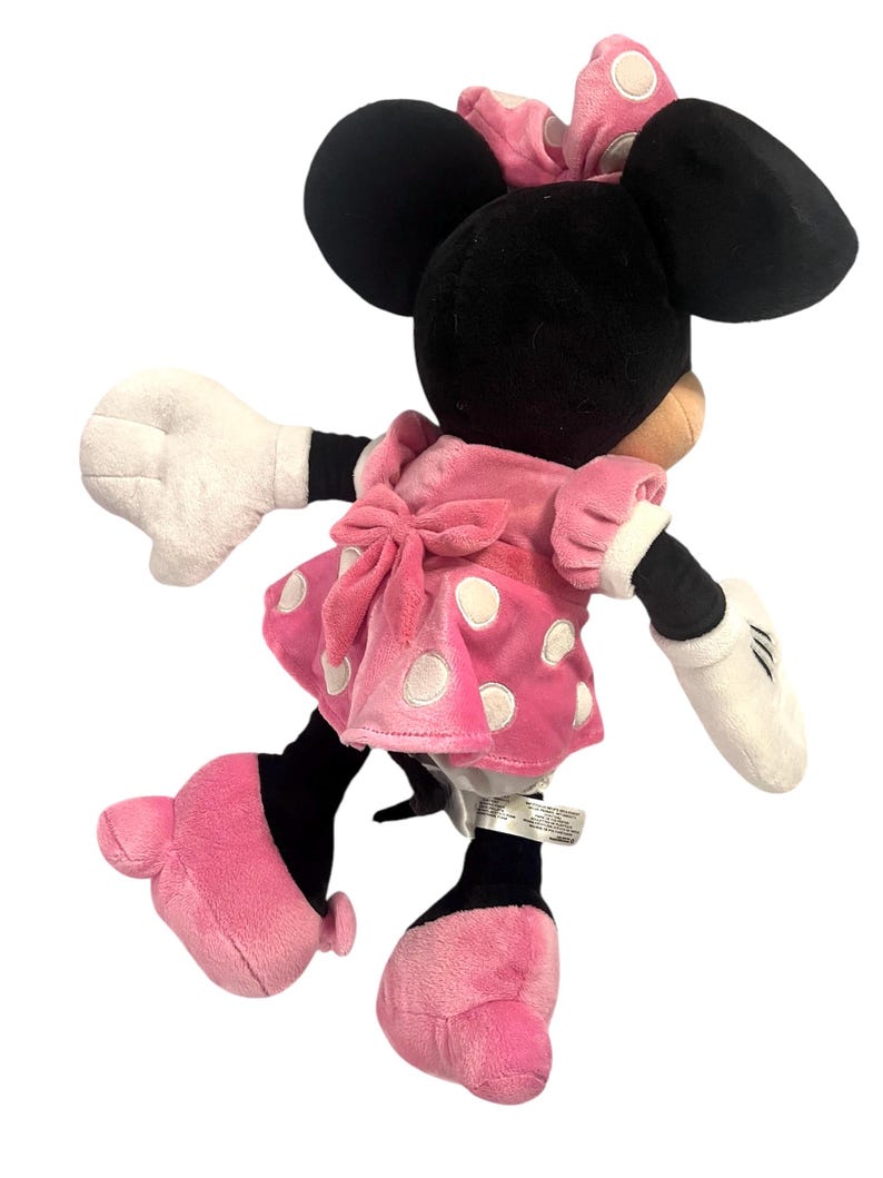 May include: A pink and white polka dot dress with a white bow on the back. The dress is worn by a plush toy of Minnie Mouse, who has black ears and a black bow on her head. The toy is standing on a white background.