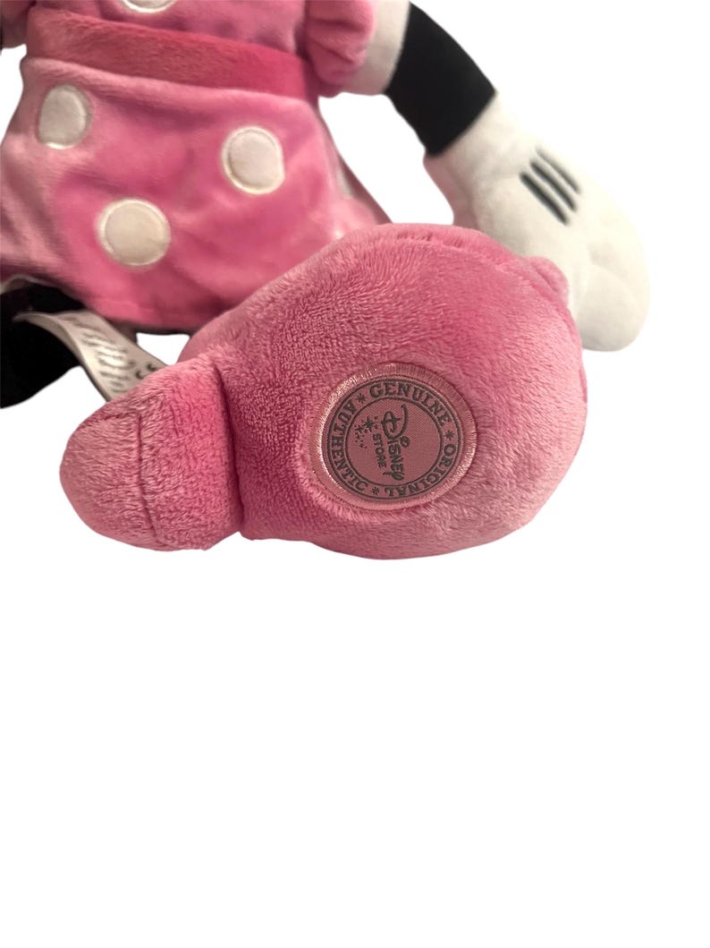 May include: Pink plush Minnie Mouse toy with white polka dots and a black bow. The toy has a white tag on its leg that says "Genuine Disney Store."