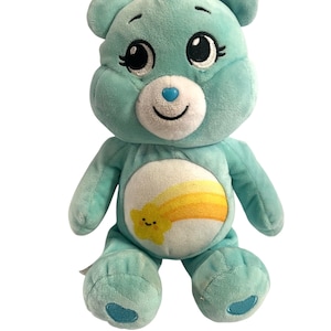 Pode incluir: Um urso de pelúcia azul claro Care Bear com um arco-íris amarelo e uma estrela sorridente no peito. O urso tem um rosto branco com olhos pretos e um nariz azul.