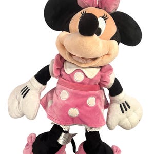May include: A pink plush Minnie Mouse toy with black ears, white gloves, and black shoes with pink bows. She is wearing a pink dress with white polka dots and a red bow around her waist.