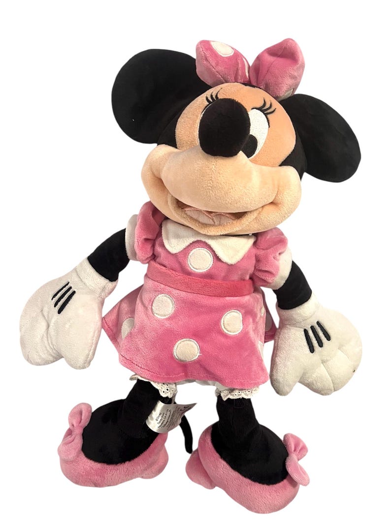 May include: A pink plush Minnie Mouse toy with black ears, white gloves, and black shoes with pink bows. She is wearing a pink dress with white polka dots and a red bow around her waist.