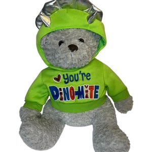 May include: A gray plush teddy bear wearing a green dinosaur costume with a silver headpiece. The costume has the text "You're Dino-mite" on the front.