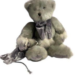 May include: A plush grey teddy bear wearing a silver velvet scarf and holding a silver velvet stocking. The bear has a white fur trim around its paws and a white fur trim around its neck.
