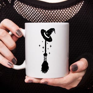 May include: White ceramic mug with a black silhouette of a witch's hat and broom with stars.