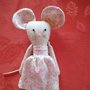 May include: A handmade plush mouse toy wearing a floral print dress. The mouse has a beige body, black eyes, and a pink nose. The dress is a light grey with pink and yellow flowers.